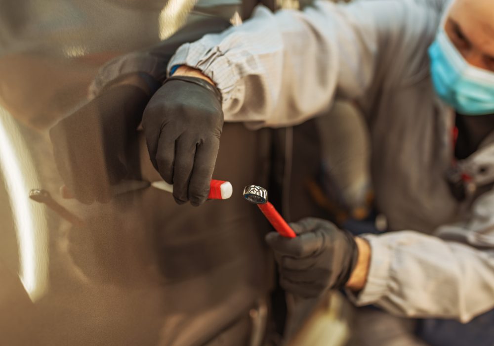 A car factory worker wearing a protective medical mask fixes a small metal defect with a hand tool.